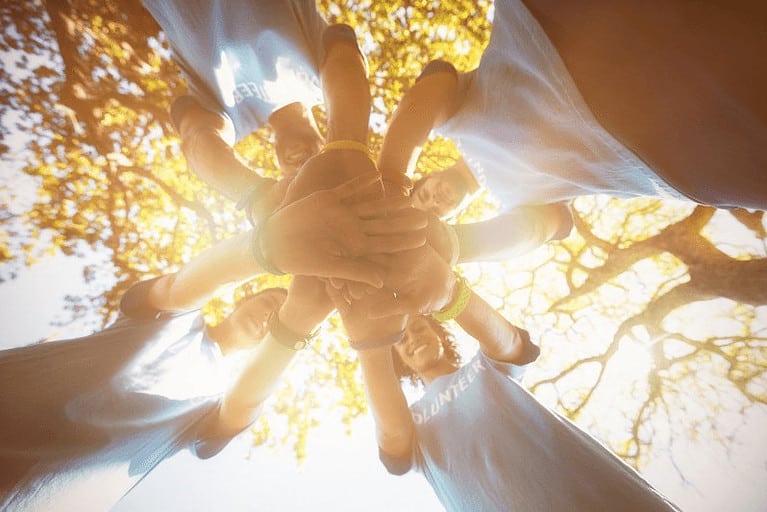 A group of volunteers stand in a circle, placing their hands together, showing they are coming together in an act of service.